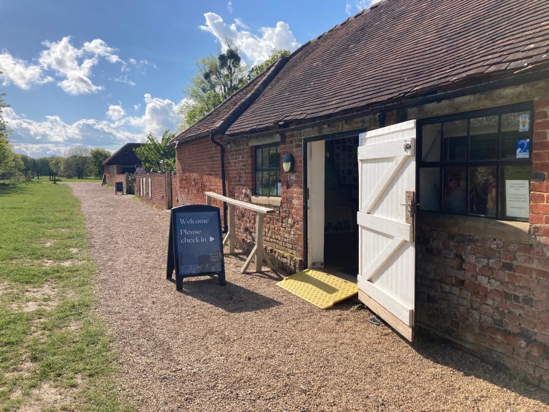 A gravel path alongside red brick buildings. There is an open door with a ramp and a sign which says Welcome Please Check in with an arrow pointing to the door.